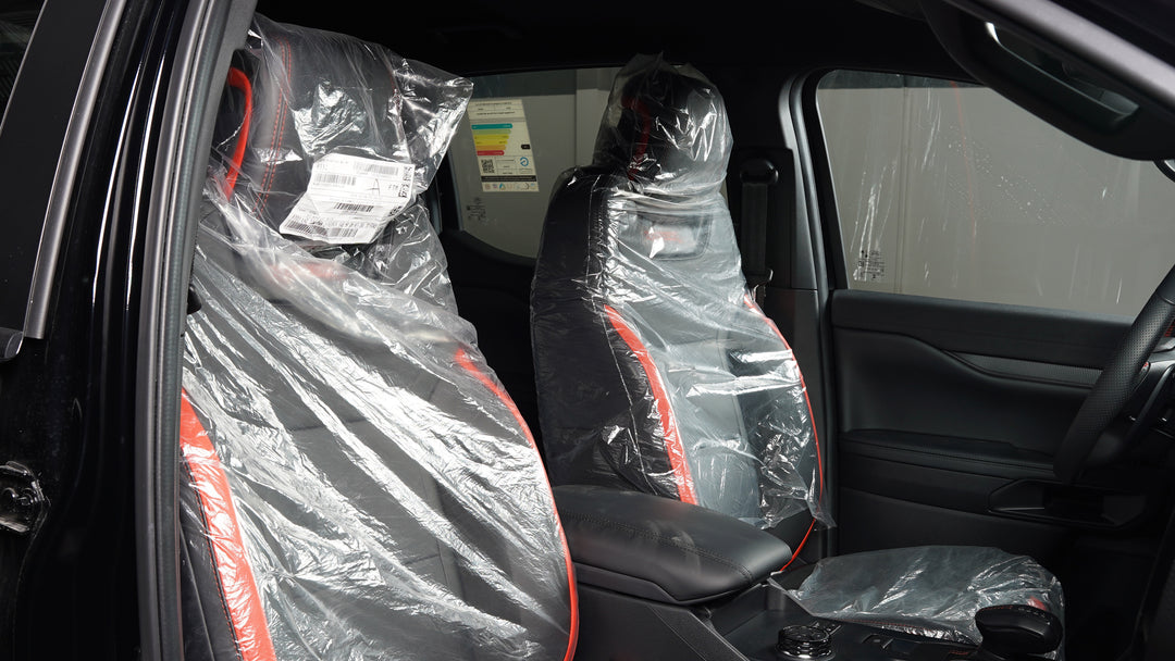 A pair of black car seat covers with red stitching, still wrapped in clear protective plastic, installed on the front seats of a vehicle interior.