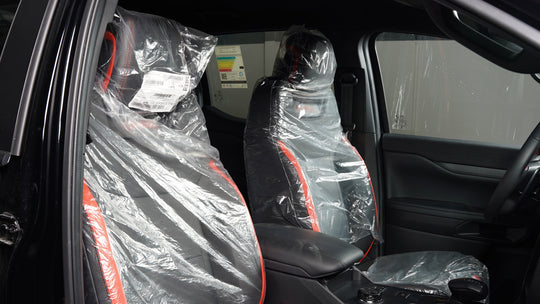 A pair of black car seat covers with red stitching, still wrapped in clear protective plastic, installed on the front seats of a vehicle interior.