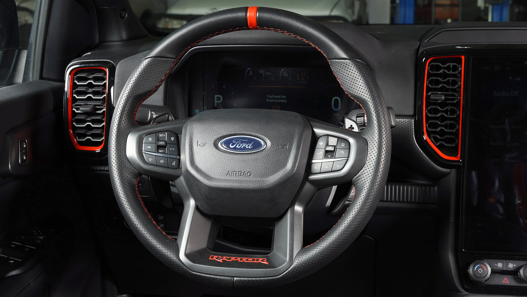 A close-up of a Ford Raptor steering wheel inside a vehicle cabin, featuring a black leather-wrapped rim with red contrast stitching, a textured center, the Ford logo, and red accent lines on the dashboard and air vents.