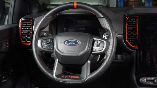 A close-up of a Ford Raptor steering wheel inside a vehicle cabin, featuring a black leather-wrapped rim with red contrast stitching, a textured center, the Ford logo, and red accent lines on the dashboard and air vents.