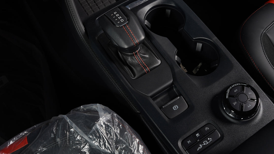Close-up of a car center console with a black gear selector, cup holders, and a round control dial, shown inside a vehicle with dark leather seats and red contrast stitching.