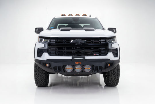 A white Chevrolet Silverado pickup truck shown from the front in a studio setting, featuring a black front bumper with integrated auxiliary lights, a large black grille, and off-road tires.